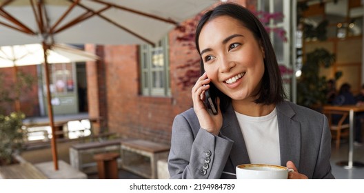 Smiling Young Adult Asian Business Woman Professional Manager, Businesswoman Entrepreneur Executive Wearing Suit Talking On Cell Phone Making Mobile Call Standing Outside Office.