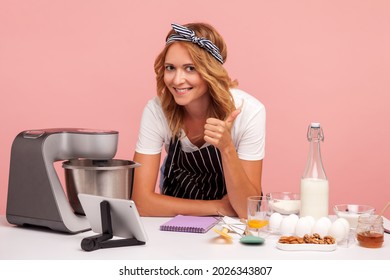 Smiling young adult baker leaning on table full of baking ingredients and food processor, looking at camera and showing thumb up, happy to do pastry. Indoor studio shot isolated on pink background. - Powered by Shutterstock