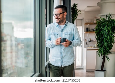Smiling young adult businessman holding drinking coffee cup in office entrepreneur looking through window while standing in modern workplace man taking a time off or taking a brake from hard work - Powered by Shutterstock