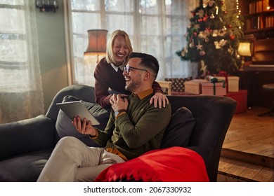 Smiling Young Adult Couple Enjoying Christmastime On Sofa And Using Digital Tablet