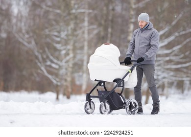 Smiling young adult father pushing white baby stroller and walking on snow covered sidewalk at park in cold winter day. Spending time with newborn and breathing fresh air. Enjoying peaceful stroll.