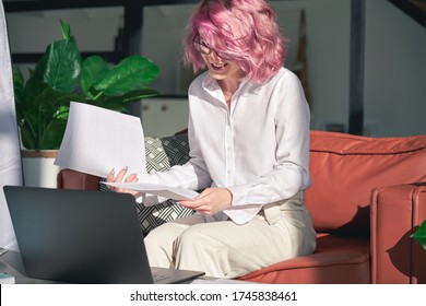 Smiling Young Adult Hipster Woman Online Teacher, Remote Worker With Pink Hair Holding Papers Using Laptop Video Conference Zoom Calling At Home Office Online Sitting On Sofa In Sunny Living Room.