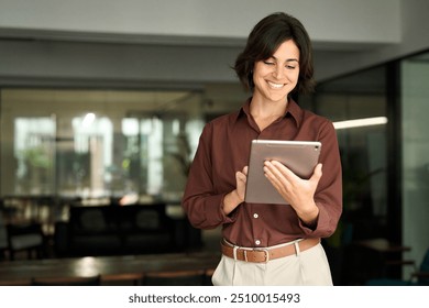 Smiling young adult Hispanic businesswoman leader or entrepreneur, company worker using tablet at work. Happy busy female professional executive holding tab computer standing in office. - Powered by Shutterstock