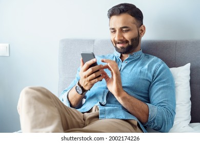 Smiling young adult indian freelance business man using mobile phone checking social media network feed or message chat sitting on bed at home. Online digital communication, rest after hard work day - Shutterstock ID 2002174226