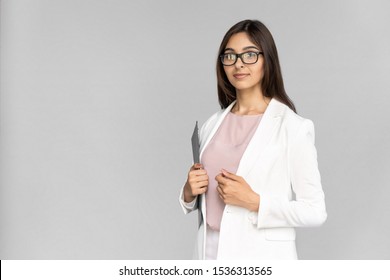 Smiling Young Adult Indian Professional Business Lady In White Formal Wear Standing Isolated On Grey Background With Copy Space. Woman Holding Laptop Computer In Hand And Looking At Camera