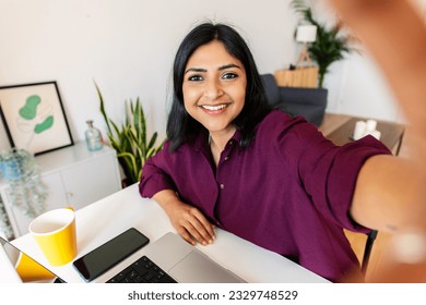 Smiling young adult indian student woman taking selfie while working on laptop at home. Face head shot portrait.