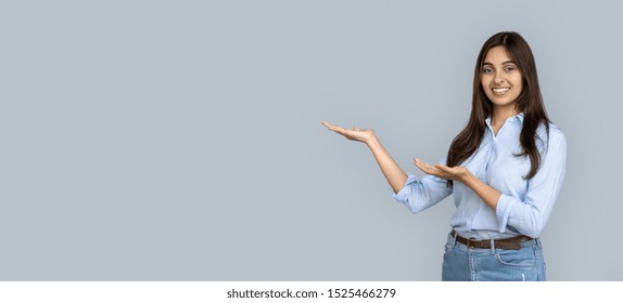 Smiling Young Adult Indian Woman Stand Isolated On Grey Background Holding Copy Space On Hands Pointing. Happy Girl Student Professional Looking At Camera Advertise Product Service Concept, Banner