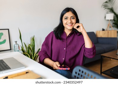 Smiling young adult indian woman holding mobile phone while working with laptop at home - Powered by Shutterstock