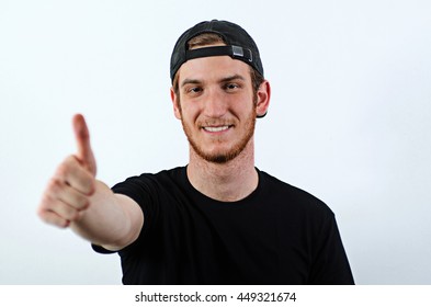 Smiling Young Adult Male In Dark T-Shirt And Baseball Hat Worn Backwards Shows His Thumbs Up