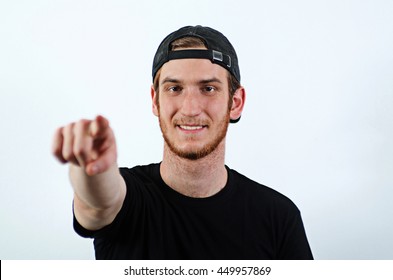 Smiling Young Adult Male In Dark T-Shirt And Baseball Hat Worn Backwards Pointing Towards The Viewer