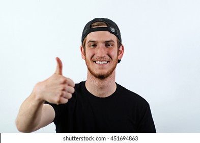 Smiling Young Adult Male In Dark T-Shirt And Baseball Hat Worn Backwards Gesturing, Showing Thumbs Up