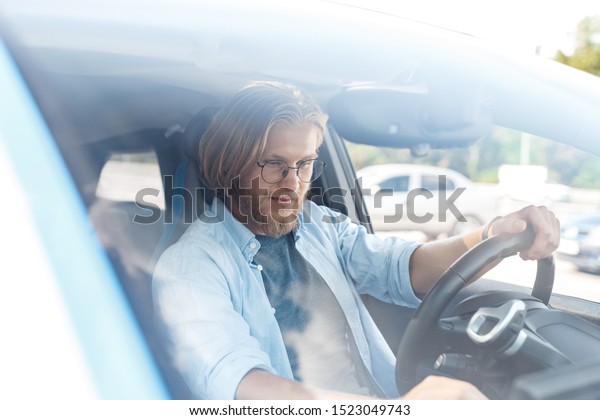 Smiling young adult man with nice calm face\
sitting in modern car, driving in city. Driver holding hand on\
steering wheel and adjusting system in\
salon