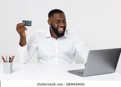 Smiling young african american business man in classic shirt posing working in office sitting at desk using laptop pc computer hold credit bank card isolated on white color background studio portrait.