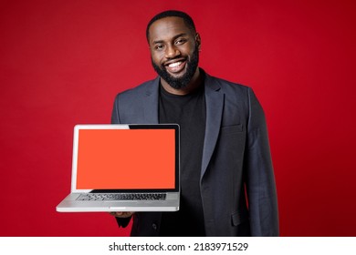 Smiling Young African American Business Man 20s In Classic Jacket Suit Standing Hold Laptop Pc Computer With Blank Empty Screen Looking Camera Isolated On Bright Red Color Background Studio Portrait