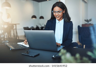 Smiling young African American businesswoman using a laptop while working at a desk in a bright modern office with colleagues in the background