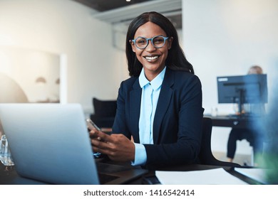 Smiling young African American businesswoman using a cellphone and working on a laptop while sitting at her desk in a bright modern office - Shutterstock ID 1416542414