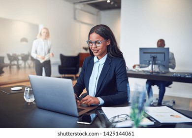 Smiling young African American businesswoman working online at her desk using a laptop in a bright modern office with colleagues in the background - Shutterstock ID 1429144061