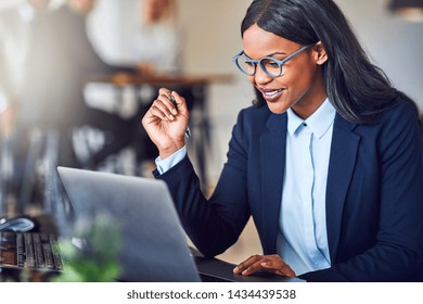 Smiling Young African American Businesswoman Working Online At Her Desk Using A Laptop In A Bright Modern Office With Colleagues In The Background