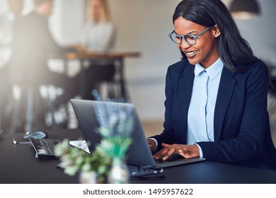 Smiling young African American businesswoman working on a laptop at her desk in a bright modern office with colleagues in the background - Powered by Shutterstock