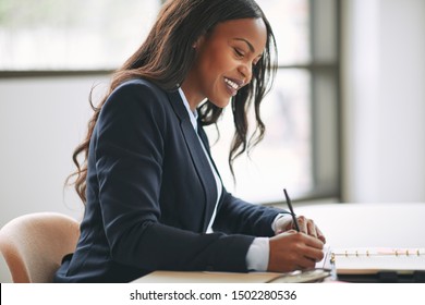 Smiling Young African American Businesswoman Writing Down Notes In Her Day Planner While Sitting At A Table In An Office