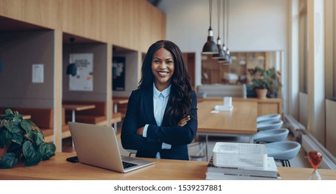 Smiling young African American businesswoman standing confidently with her arms crossed while working on a laptop in an office lounge