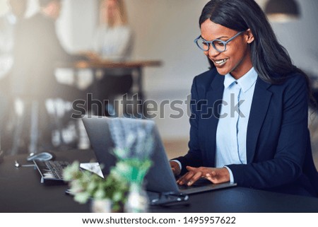 Smiling young African American businesswoman working on a laptop at her desk in a bright modern office with colleagues in the background