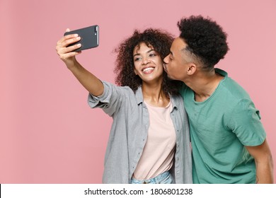 Smiling young african american couple friends man woman in gray green casual clothes posing doing selfie shot on mobile phone kissing in cheek isolated on pastel pink color background studio portrait