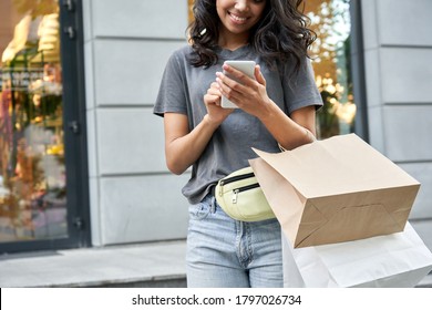 Smiling Young African American Ethnic Girl Shopper Customer Standing On City Street Holding Shopping Bags And Smart Cell Phone Using Mobile Fashion Sale Ecommerce App Near Shop Mall Store, Closeup.