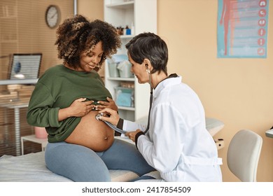 Smiling young African American expectant mother sitting on medical table in clinic while female obstetrician using stethoscope listening baby - Powered by Shutterstock