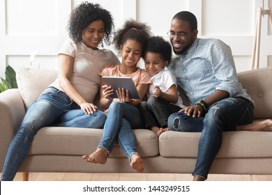 Smiling Young African American Family Sit On Couch With Little Kids Watch Cartoons On Tablet, Happy Black Parents Have Fun Relax With Children On Sofa Enjoy Video On Pad Spending Time At Home Together