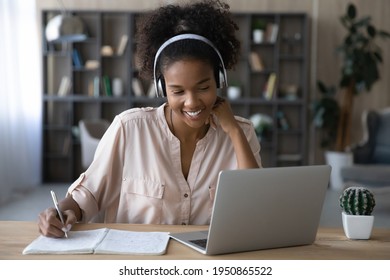 Smiling young African American female in headphones write look at laptop screen study online on gadget. Happy millennial biracial female in earphones work distant on computer. Education concept.