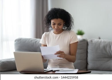 Smiling Young African American Girl Student Reading Paper Post Mail Letter Of Advice Bank Statement Invoice Sit On Sofa, Happy Black Woman Holding Bill Document Notification Doing Paperwork At Home