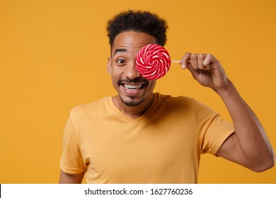 Smiling young african american guy in casual t-shirt posing isolated on yellow orange background studio portrait. People emotions lifestyle concept. Mock up copy space. Cover eye with round lollipop - Shutterstock ID 1627760236