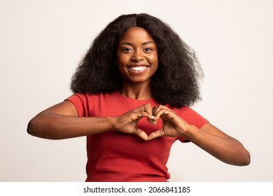 Smiling Young African American Lady Showing Heart Gesture With Hands Near Chest, Happy Black Millennial Lady Making Love Sign At Camera While Posing Over White Studio Background, Copy Space