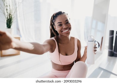 Smiling young African American lady with bottle of water taking selfie on break from home workout. Positive black woman making photo of herself during domestic training, indoors - Powered by Shutterstock