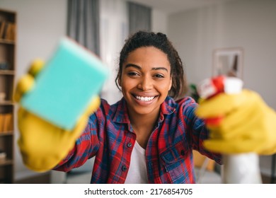 Smiling young african american lady in rubber gloves with spray and sponge wipes camera at modern kitchen interior. Perfectionism, cleaning, housework, hygiene and housewife have fun at home, covid-19 - Powered by Shutterstock