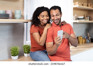 Smiling Young African American Male And Female In Same T-shirts Have Video Call On Smartphone Or Watch Video In Cozy Kitchen Interior. Online Chat, Communication Remotely At Home, Social Distance