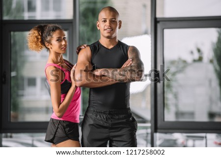 smiling young african american male athlete posing with crossed arms while his girlfriend standing near at gym