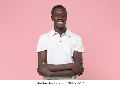 Smiling Young African American Man Guy In White Polo Shirt Posing Isolated On Pastel Pink Wall Background Studio Portrait. People Emotions Lifestyle Concept. Mock Up Copy Space. Holding Hands Crossed