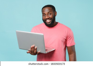 Smiling young african american man 20s in casual pink t-shirt isolated on pastel blue wall background studio portrait. People lifestyle concept. Mock up copy space. Working on laptop pc computer