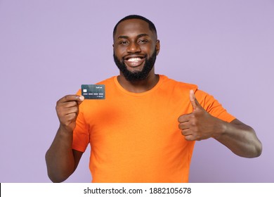 Smiling young african american man 20s wearing basic casual orange blank empty t-shirt standing hold credit bank card showing thumb up isolated on pastel violet colour background studio portrait