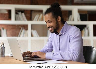 Smiling Young African American Man Look At Laptop Screen Browsing Internet On Gadget. Happy Mixed Race Ethnicity Male Employee Work Online On Computer At Home Office. Digital, Technology Concept.