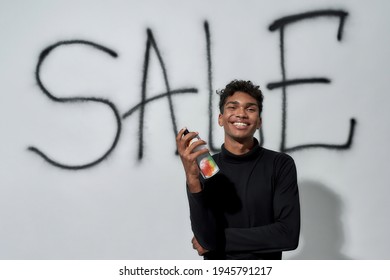 Smiling Young African American Man Holding Paint Sprayer While Standing Near Graffiti On White Wall Background And Looking At Camera. Discount And Sale Concept