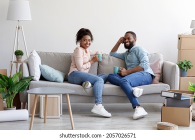 Smiling Young African American Man And Woman Relax On Couch, Drinking Coffee In Room With Cardboard Boxes With Stuff And Plants. Relocation, Housing Improvement, Life Changes, First Own House Purchase