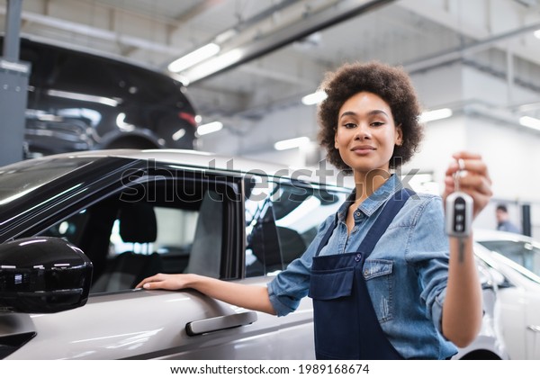 smiling young african american mechanic in\
overalls holding key near car in\
garage