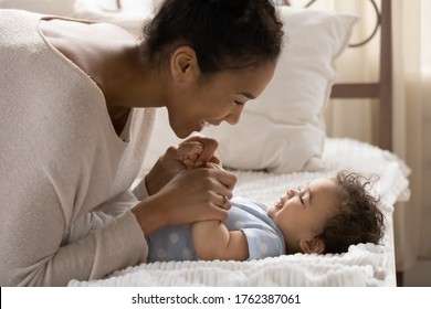 Smiling young African American mother playing with cute little daughter in bedroom close up, caring beautiful mum and toddler girl lying on cozy bed, holding hands, having fun, childcare concept - Powered by Shutterstock