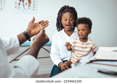 Smiling Young African American Mother With Small 2s Boy Child Son Have Consultation With Caring Male Pediatrician In Private Hospital Or Clinic. Attentive Male Doctor Support Cheer Little Kid Patient.