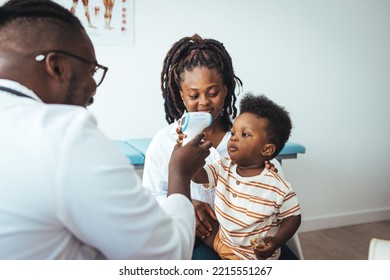 Smiling Young African American Mother With Small 2s Boy Child Son Have Consultation With Caring Male Pediatrician In Private Hospital Or Clinic. Doctor Measuring Temperature Of Boy 