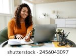 Smiling young African American woman sitting at desk working on laptop taking notes in notebook, happy millennial female studying online, watching webinar using computer and writing check list