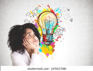 Smiling young African American woman in white shirt looking at colorful lightbulb sketch drawn on concrete wall. Concept of good idea